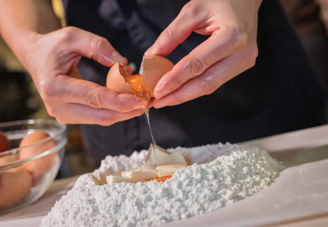 Primo piano di mani che rompono un uovo fresco nella farina per preparare l'impasto della pasta fatta in casa.