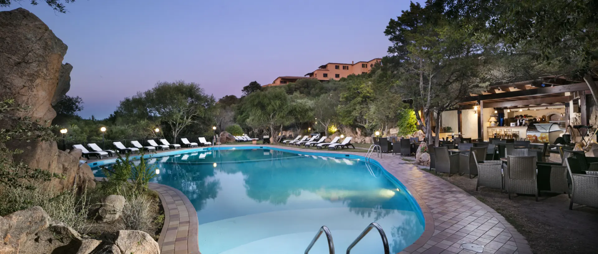 Panoramica della piscina al crepuscolo con lettini prendisole e vista sull'edificio principale del Myo Hotel Rocce Sarde.