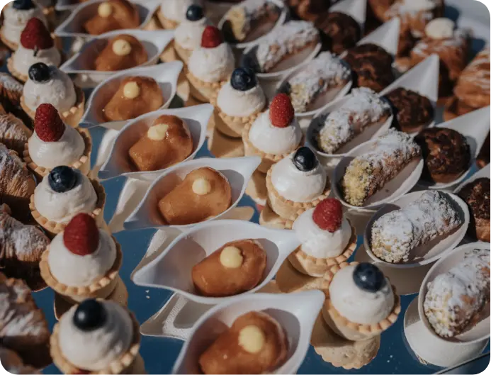 Ricco buffet di dolci monoporzione che include babà al rum, tartellette con crema e frutti di bosco, e piccoli cannoli siciliani.