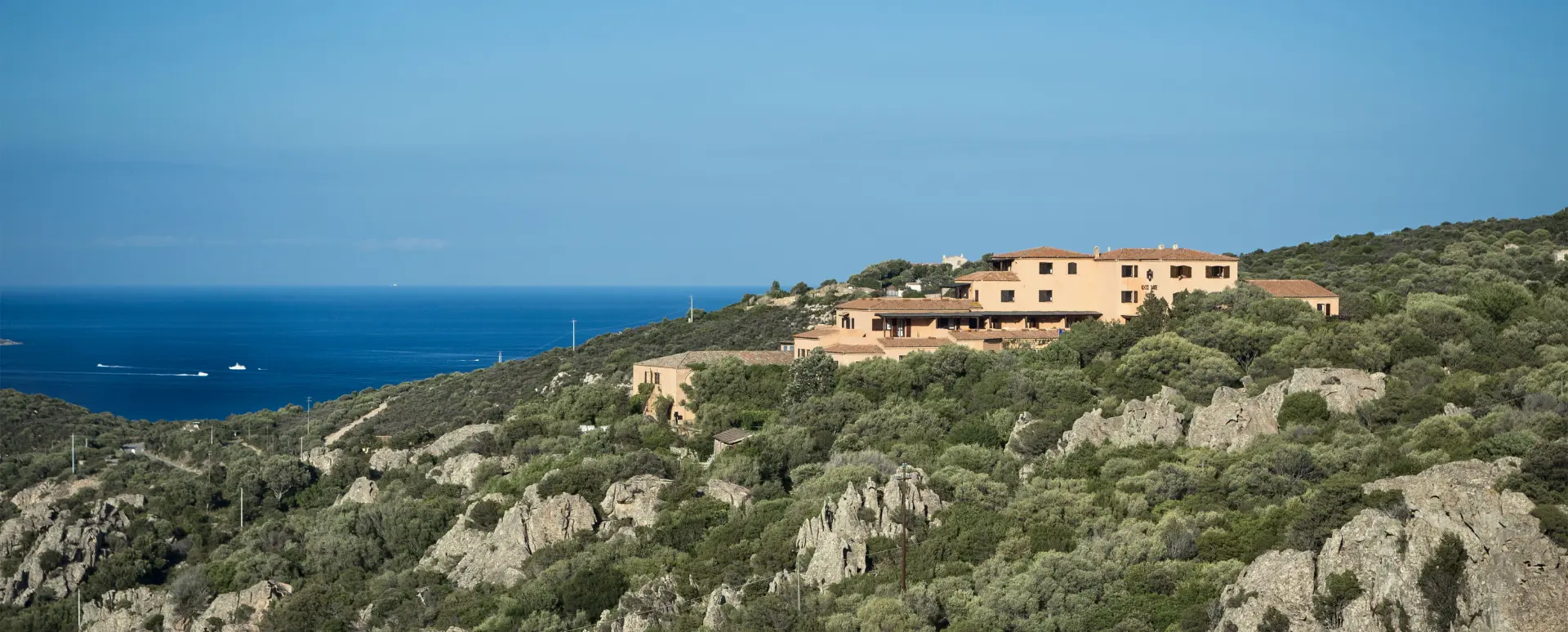 Vista laterale dell'albergo sulla collina rocciosa, immerso nella macchia mediterranea con il mare azzurro sullo sfondo.