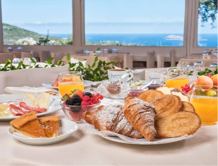 Tavola per la colazione con brioches, frutta fresca, succhi e salumi con una vista panoramica sul mare.