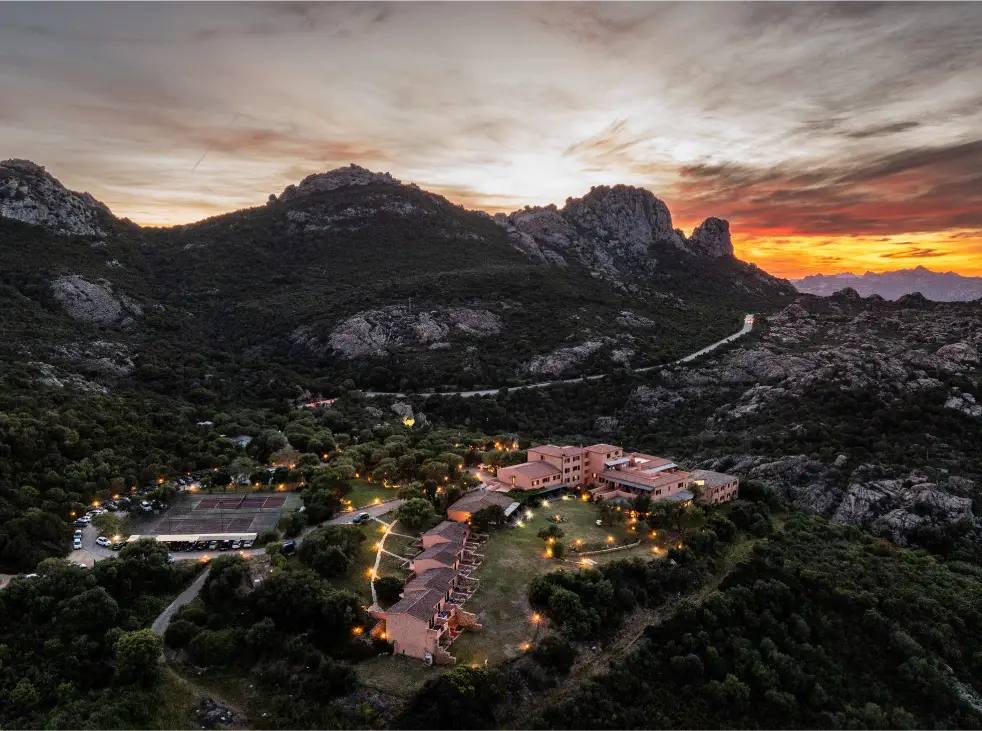 Hotel Rocce Sarde al crepuscolo, illuminato, circondato da montagne e vegetazione.