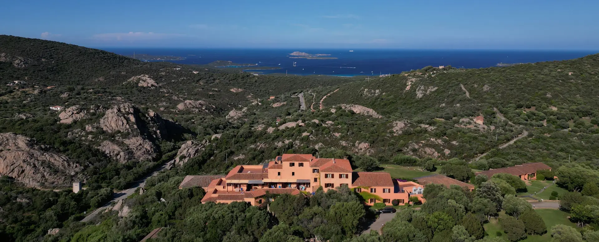 Vista panoramica diurna dell'Hotel Rocce Sarde immerso nel verde con il mare e isole sullo sfondo.